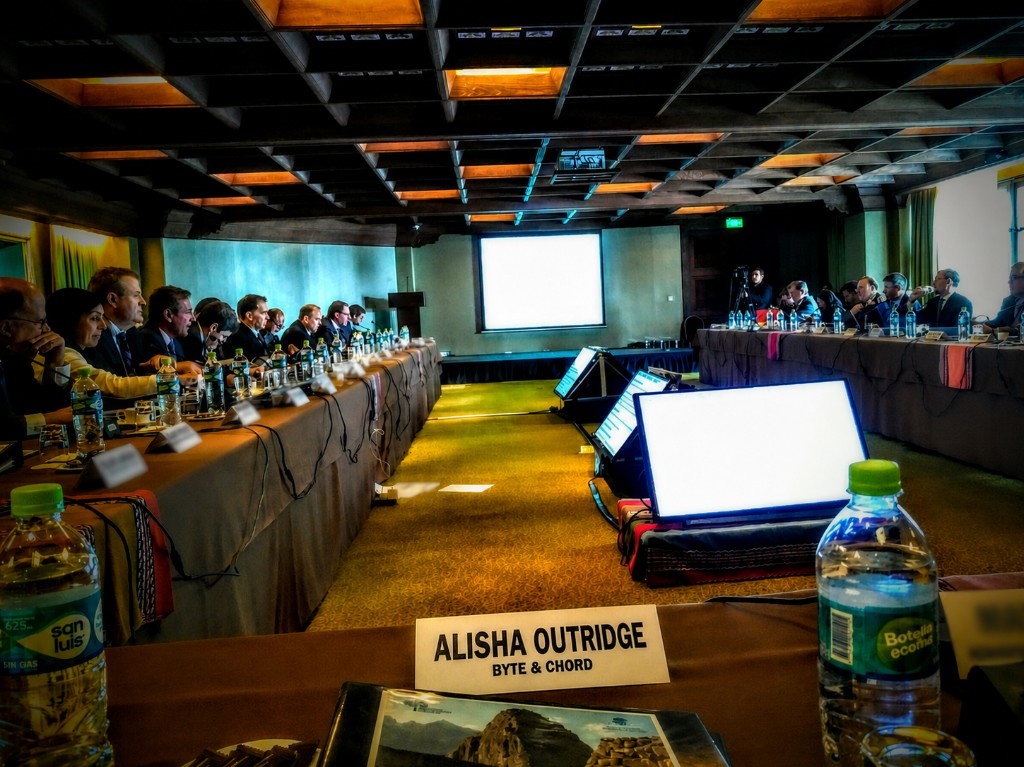 Boardroom at a private executive offsite with leadership teams seated at long tables, Alisha Outridge's Byte&Chord name card in the foreground